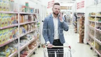 穿着白色衬衫的年轻微笑男子正在智能手机上交谈，并在超市的货架之间行走着商品。